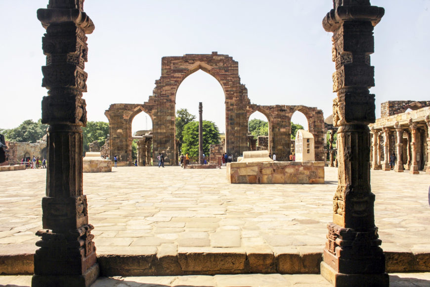 El patio de la mezquita Qutb, c. 1192, complejo arqueológico Qutb, Delhi (foto: Indrajit Das, CC BY-SA 4.0). En primer plano se encuentran pilares de la pasarela con columnas y al fondo se encuentra un pilar de hierro del siglo IV al V y la pantalla arqueada y sala de oración de la mezquita.