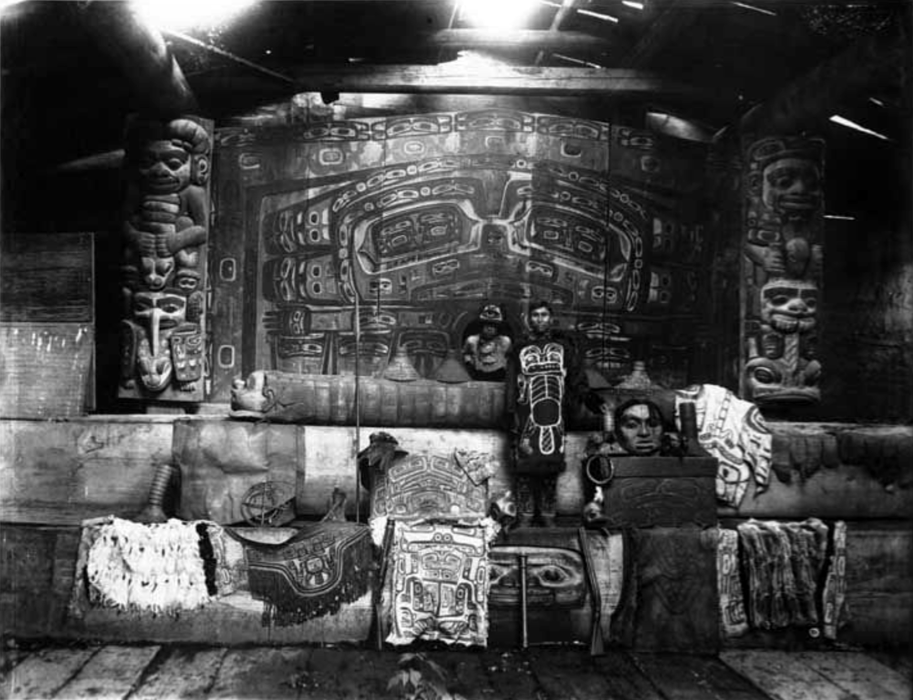 Interior of Yáay Hít (Whale House) of the Tlingit Ghaaanaxhteidí clan, Klukwan, Alaska, c. 1895. The photo shows the at.óow (Tlingit clan property) of the Ghaaanaxhteidí clan, including the Séew Xh'éen (Rain Screen), Wormwood house post (left), Strong Man house post (right), and tunics, bentwood boxes, and clan hats. Photo by Winter and Pond (Alaska State Libraries)