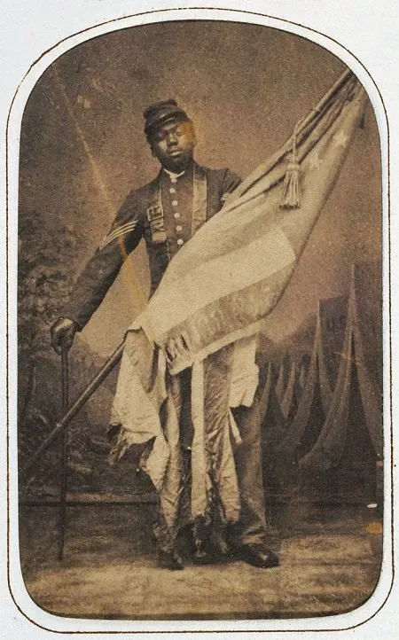 Sergeant William Carney holding the American Flag, c. 1864.  John Ritchie, Carte-de-viste album of the 54th Massachusetts Infantry Regiment, c. 1864 (<a href="https://edan.si.edu/slideshow/viewer/?damspath=/Public_Sets/NMAAHC/NMAAHC_Slideshows/2014_115_8" target="_blank" rel="noopener">National Museum of African American History and Culture</a>)