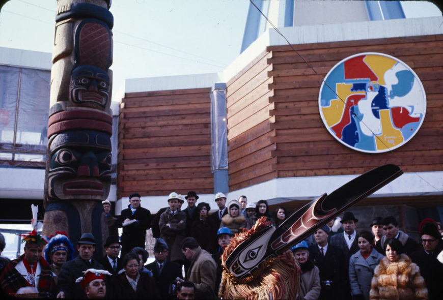 Detail of totem pole by Henry and Tony Hunt and Alex Janvier’s The Unpredictable East (Beaver Crossing, Indian Colours) at the Indians of Canada Pavilion, with Harry Hunt performing the Raven Dance in the foreground. 1967. e011194636, Library and Archives Canada / Canadian Corporation for the 1967 World Exhibition