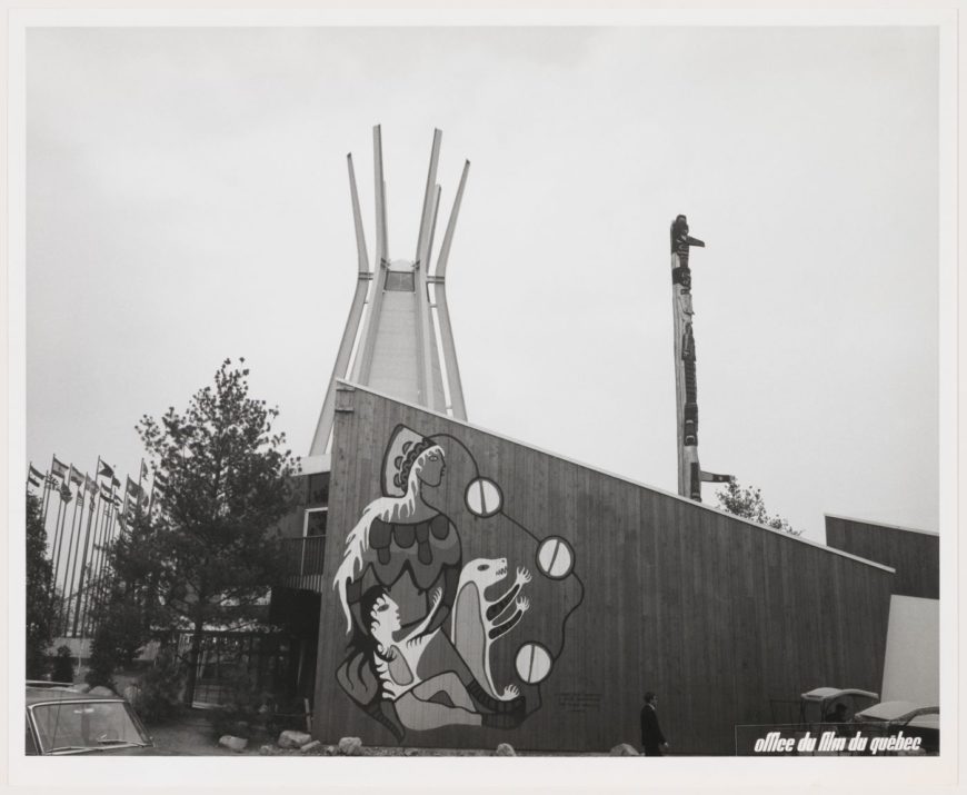 A black-and-white photograph of Anishinaabe artist Norval Morrisseau’s mural at the Indians of Canada Pavilion. The mural, painted directly on the vertical cedar boards of the building, depicts the Earth Mother with long, white hair, a head covering, and exposed breasts, along with a child with long black hair, and a white and yellow bear cub. A series of circular forms linked with black lines connects the figures to the ground plane. The “tipi” and totem pole are visible in the background. Norval Morrisseau, Earth Mother with Her Children, 1967