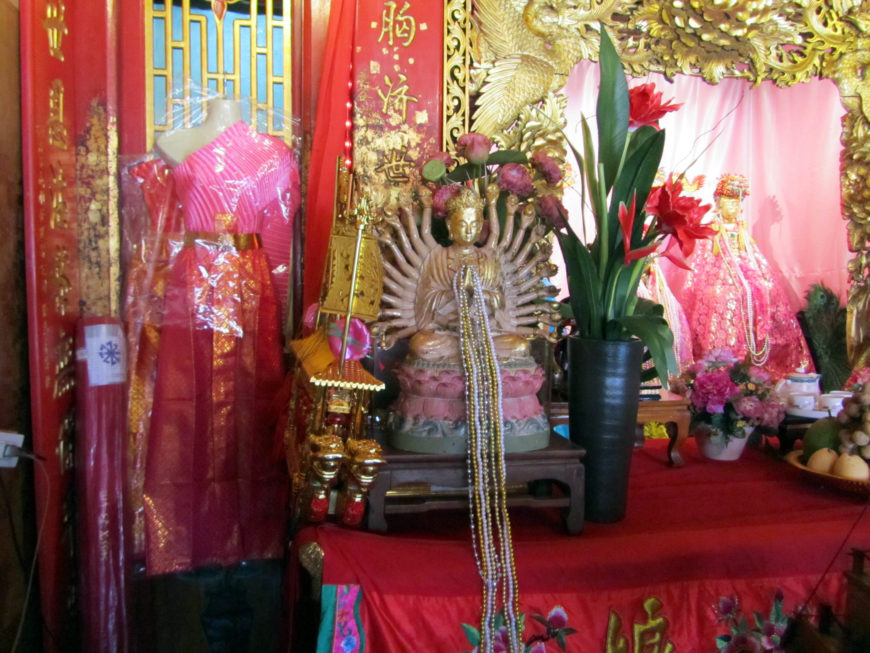 A corner of the shrine to Chao Mae Soi Dokmak at Wat Phanan Choeng, Ayutthaya. The figure of the princess is in the framed niche to the right. The statue of thousand-armed Guanyin in the foreground bears pearl necklaces that, together with pretty fabric and fancy outfits, are popular offerings to the spirit (photo by the author).