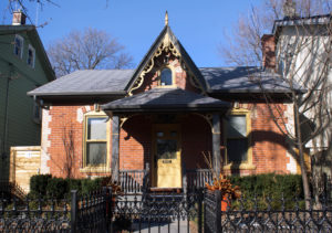 A red brick house with yellow brick details. The door and window frames, and the bargeboard and finial are painted yellow. There is a small, raised porch framing the entrance.
