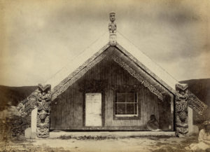 Front of a Māori community meeting house with carved end posts on either side of the veranda, maihi (carved barge-boards), raparapa (projecting boards at the end of the maihi), tekoteko (figurative carving at the front of the apex of the roof), koruru (carved mask depicting the ancestor the house is named for, placed below thetekoteko), and pare (carved lintel above doorway window). Josiah Martin, Exterior of a Māori marae (community meeting house), Hinemihi, in the village of Te Wairoa, 1881, albumen print, 15.3 x 20.5 cm (© The Trustees of the British Museum, London)