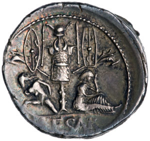Issued by Julius Caesar, the coin depicts a trophy installation made of seized carnykes (Celtic trumpets with animal heads), shields, a torc, helmet, and armor. Beneath are a clothed woman and a nude, bearded man with bound hands, and the worn letters of Caesar’s name. Roman denarius, 46 B.C.E. silver, 1.78 cm diameter (American Numismatic Society, New York)