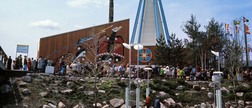 Color photograph of the exterior of the Indians of Canada Pavilion at Expo 67. The pavilion is in the form of an abstracted tipi, with bright blue steel beams serving as “poles” and white vinyl cladding. Other elements of the pavilion are clad in cedar boards, with artwork hung or painted directly on the surface. A totem pole emerges from behind part of the pavilion and a crowd of visitors is lined up in front. Indians of Canada Pavilion, Expo 67, Montreal, 1967 (Library and Archives Canada / Canadian Corporation for the 1967 World Exhibition)