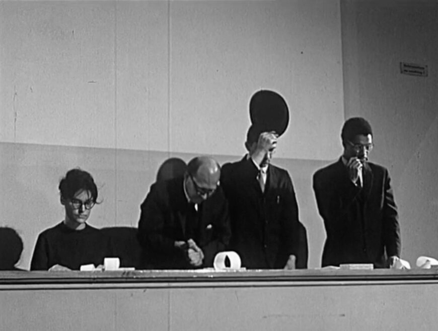 Still from digitized German TV news report on a Fluxus festival held at the Museum Wiesbaden, Germany, in September 1962. George Maciunas, Dick Higgins, Nam June Paik, Alison Knowles, Emmett Williams, George Maciunas, Benjamin Patterson performing in George Maciunas, In Memoriam to Adriano Olivetti, performed at the Fluxus Internationale Festspiele Neuester Musik, Städtisches Museum, Wiesbaden, September 8, 1962 (watch the film here: The Scores Project, Getty Research Institute, Los Angeles) © Hessischer Rundfunk