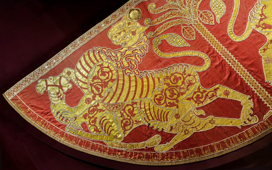 Lion, camel, and inscription (detail), Mantle of Roger II, 1133/34 (Norman Sicily, Italy), textiles, patterned samite (kermes dye), gold and silk embroidery, pearls, gold with cellular enamel, rubies, spinels, sapphires, garnets, glass, tablet weave, 146 x 345 cm (Kunsthistorisches Museum, Vienna, XIII 14; photo: Steven Zucker, CC BY-NC-SA 2.0)